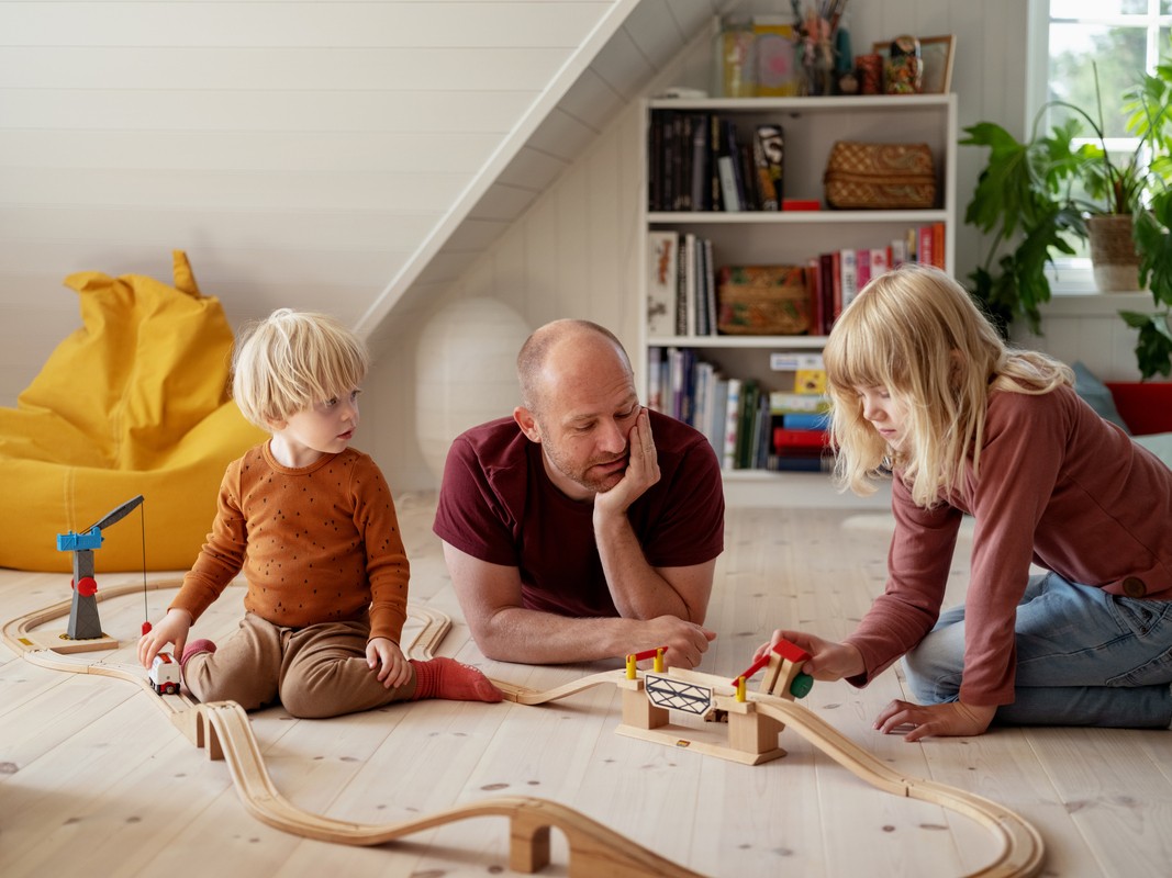 mann med datter og sønn leker med togbane på gulvet i huset sitt